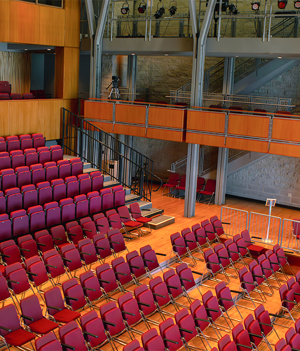 Interior of an auditorium with rows of red seats and a wood stage, with an upper balcony and lighting rig above.